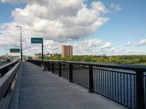 Mikey's bicycle is leaning against a railing on a bridge. Above his bicycle is a sign that reads "Quebec".