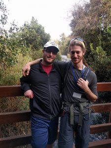 Mikey and D smile on a bridge over a river. There is a lot of green overgrowth all around.