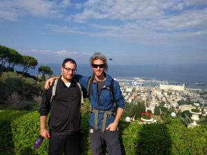 D & Mikey stand in front of a hedge on a hill overlooking a garden with a golden-topped dome. Behind that is a city and then a sea stretching to the horizon.