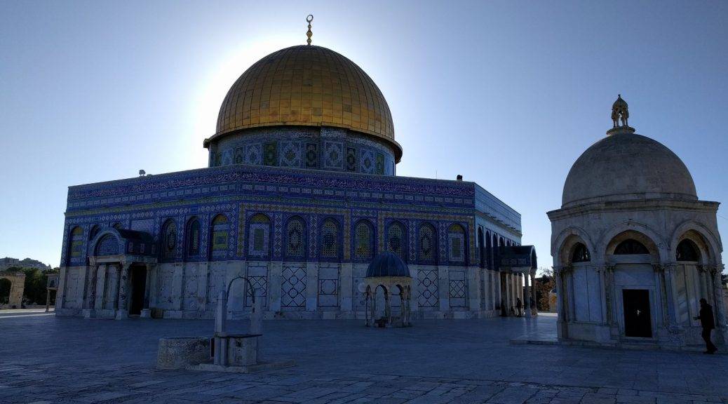 The sun low in the sky, rising in the background. It is near-symmetrically silhouetting the golden top of the Dome of the Rock atop the Temple Mount in the old city of Jerusalem (Al-Quds).