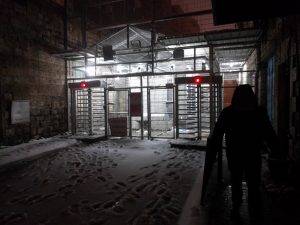 A black figure stands in the right of the shot. He is silhouetted by the well-lit cage behind him--a fenced-off military checkpoint. The street leading-up to the checkpoint is covered in white snow, and there are footprints leading to the turnstiles in & out of the checkpoint. The turnstiles both display a red X over them.